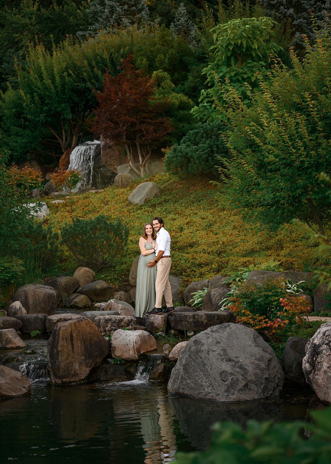 Couple embracing on rocks by a pond, surrounded by lush greenery and a small waterfall.
