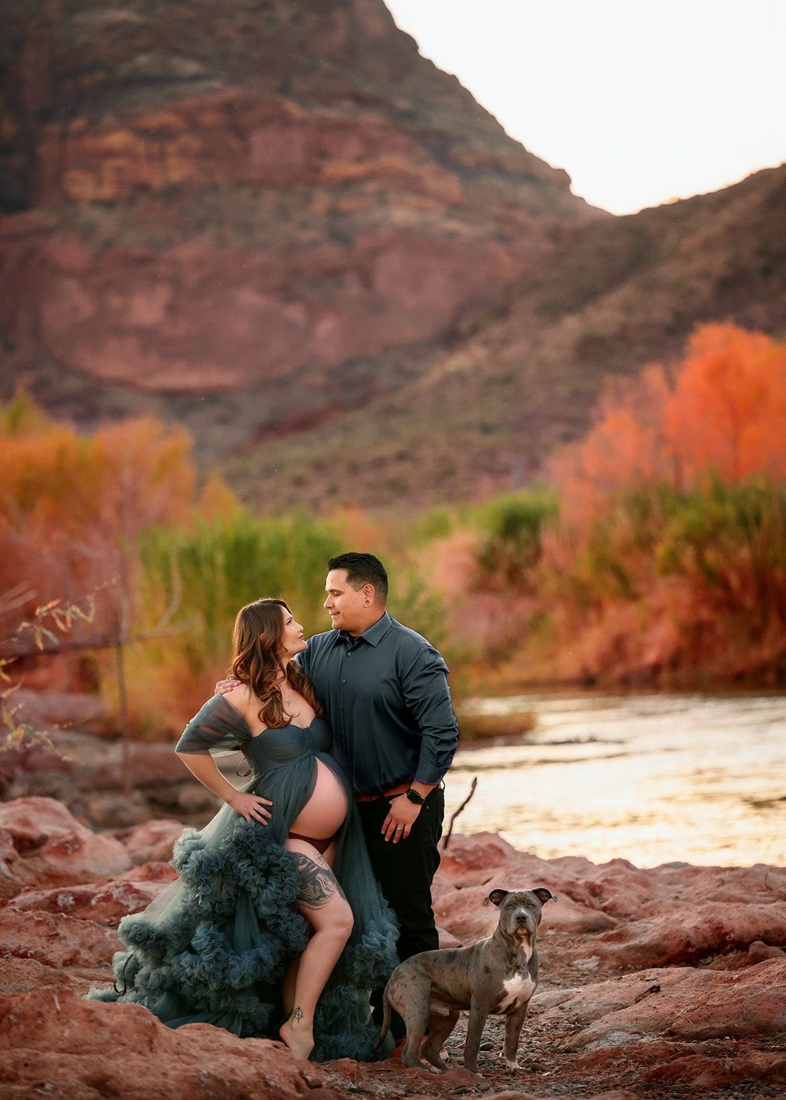Couple posing by a river with a dog, red rocks and a mountain backdrop at sunset