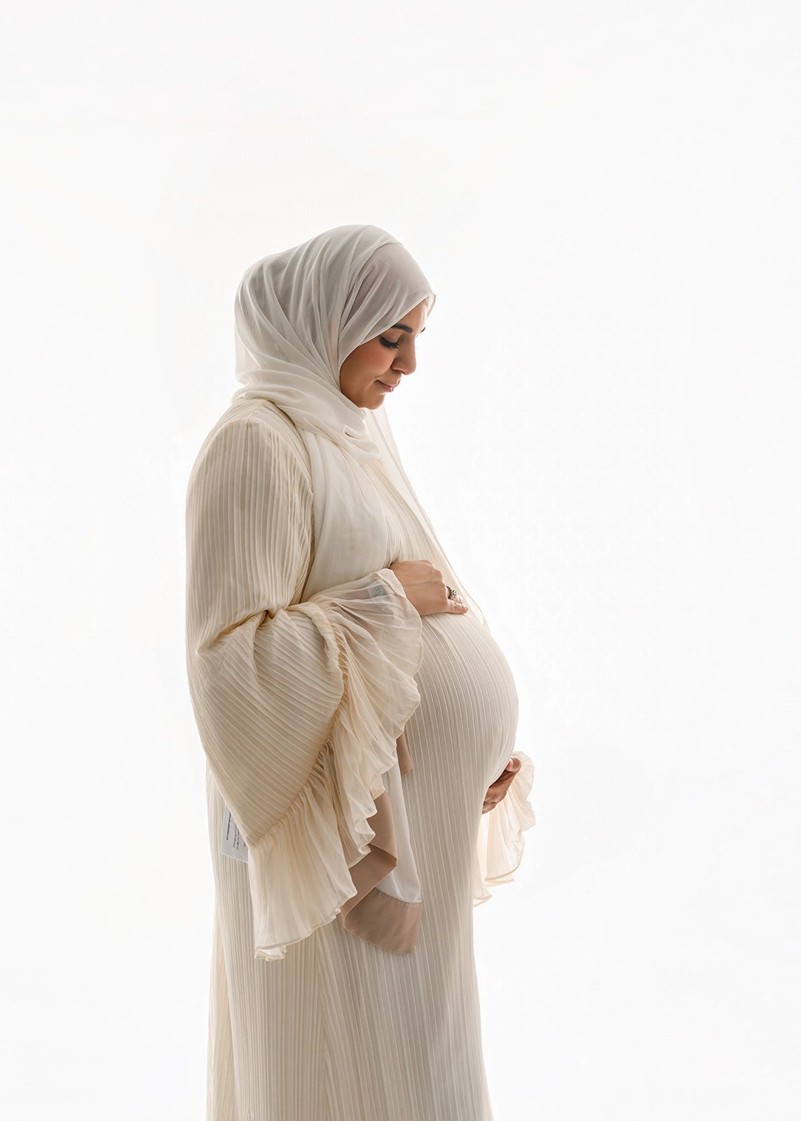 Pregnant person in a cream dress and headscarf, standing in profile against a white background, hands on belly