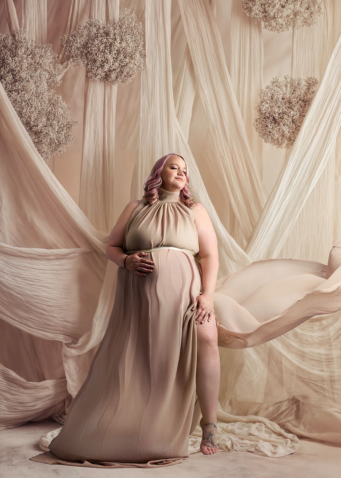 Woman in a flowing beige gown posing in a soft draped studio set with floral wall decor