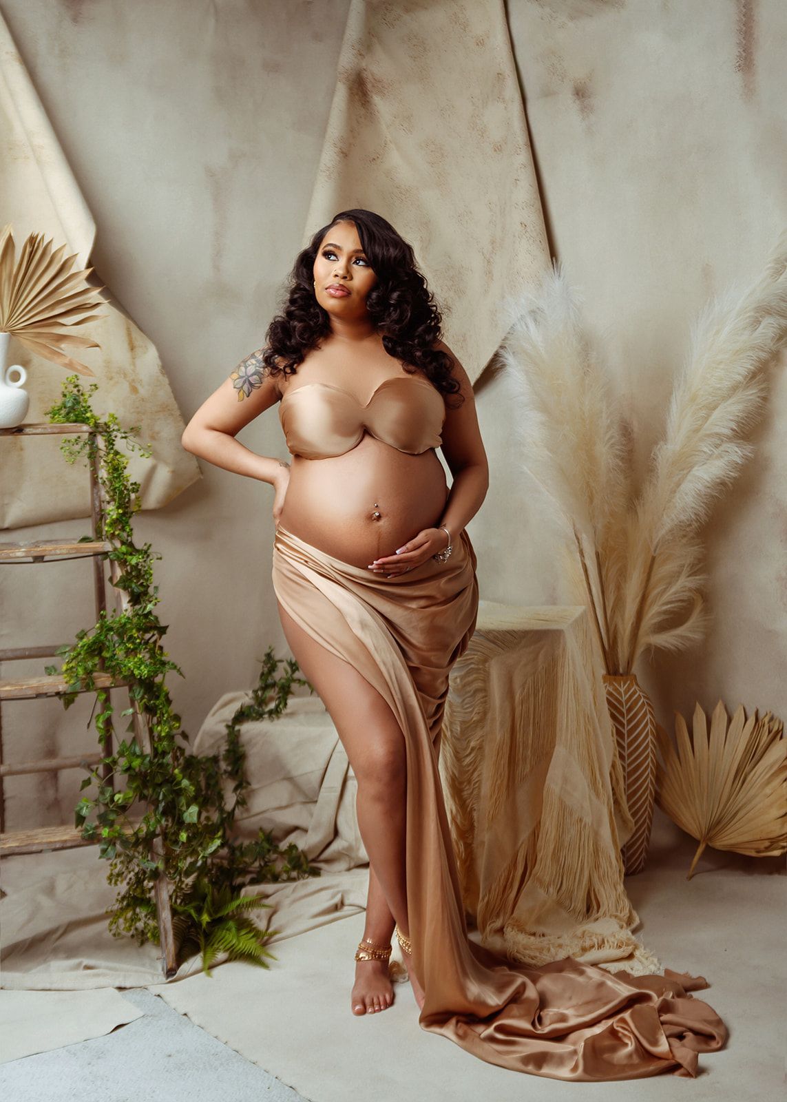 Pregnant model in a flowing beige gown posing in a studio with dried grasses and neutral drapery.