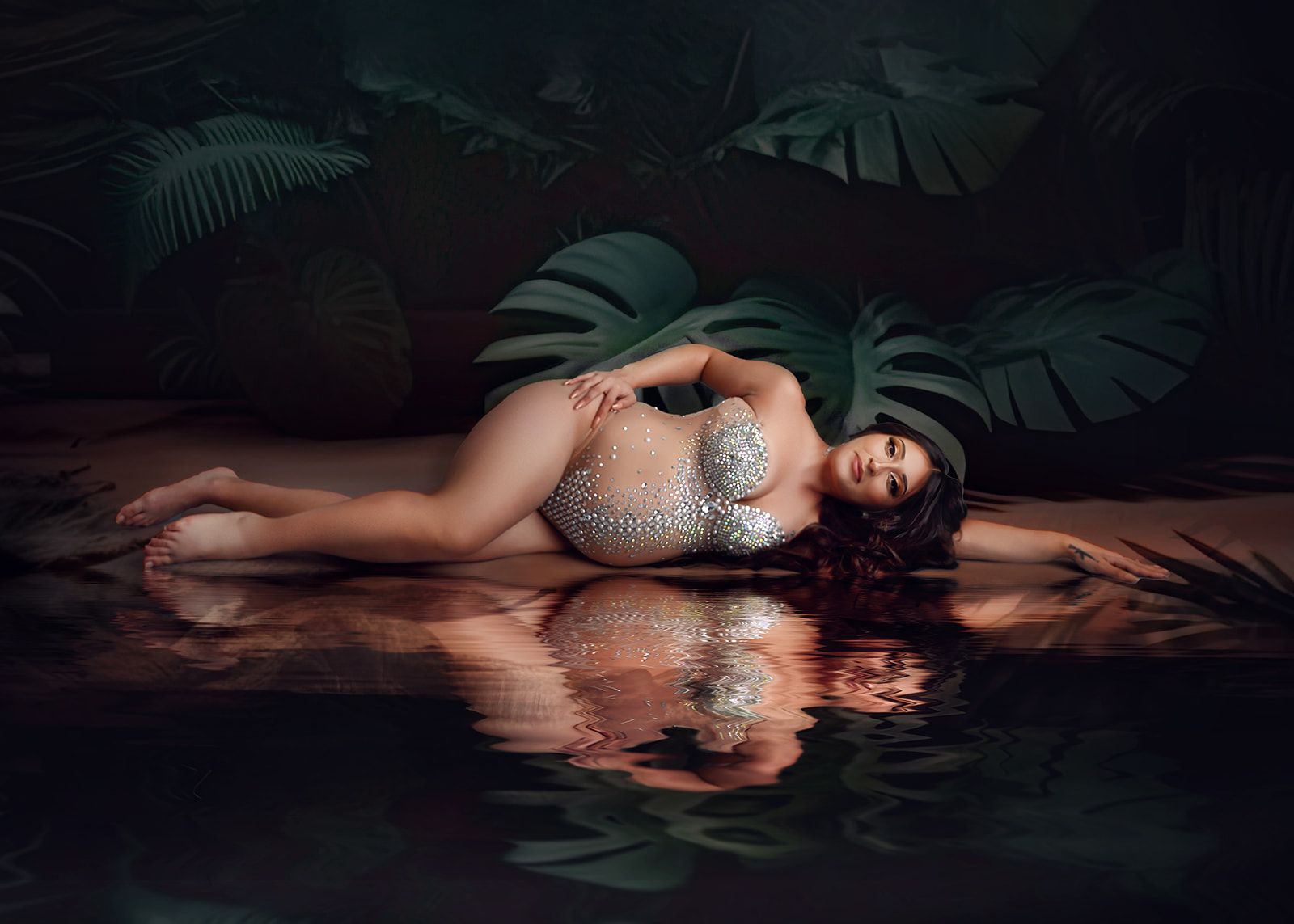 Woman lying on her side in a reflective pool, wearing a sparkling silver outfit amid dark tropical leaves