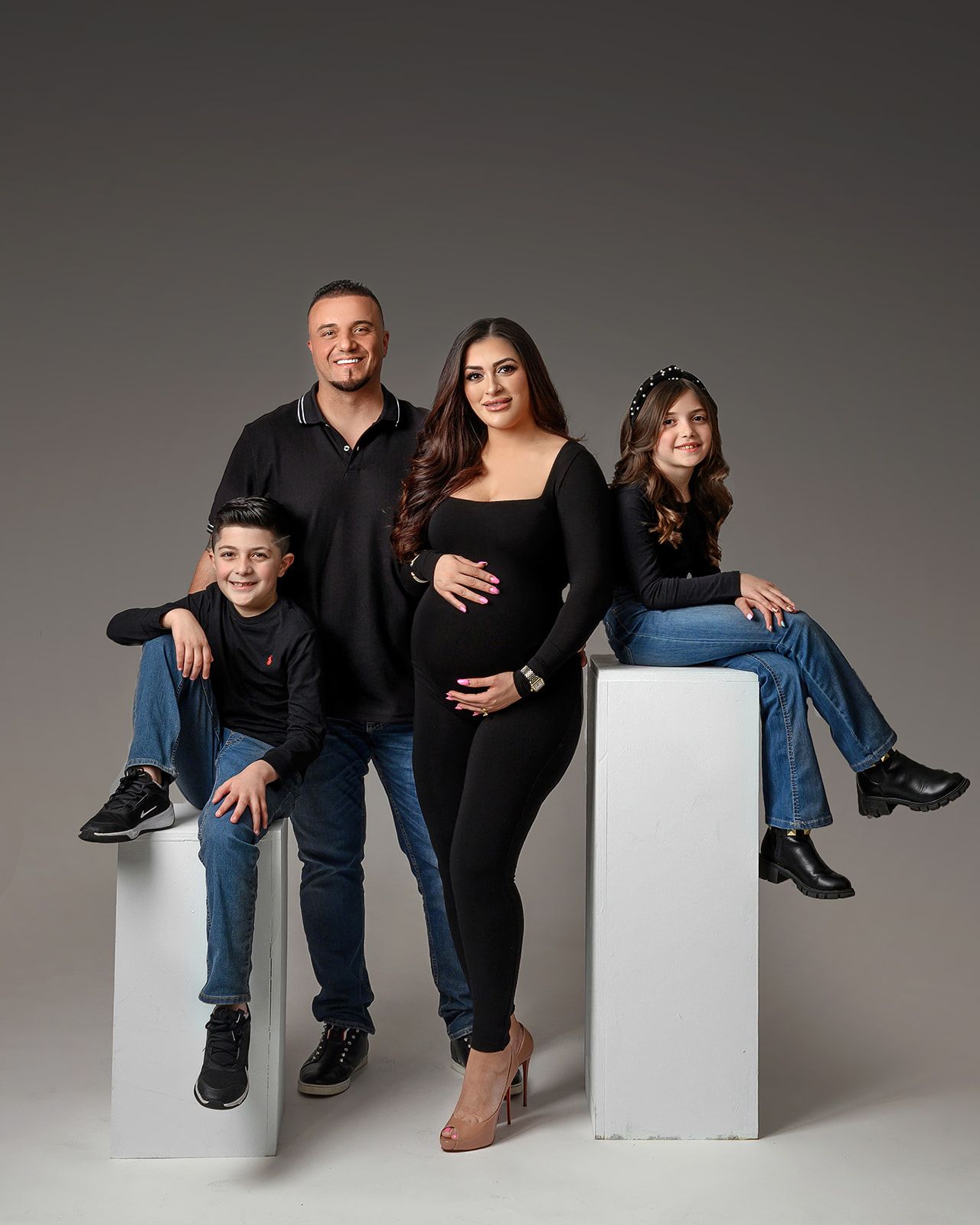 Four people posing in black and denim outfits against a gray studio background, seated on white blocks.