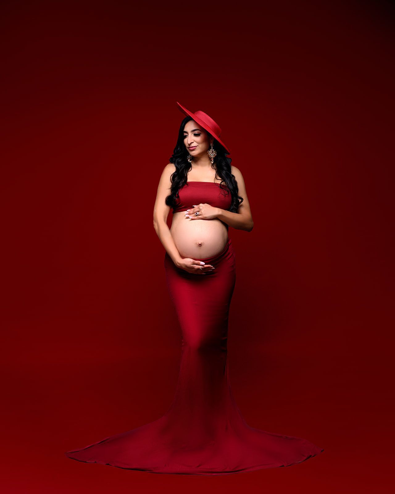 Pregnant woman in a red gown and veil, standing against a dark red background.