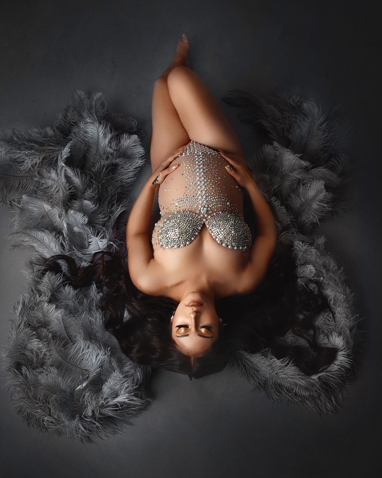 A woman lies on dark fabric, wearing a sparkling silver bodysuit, framed by black feathery textures.