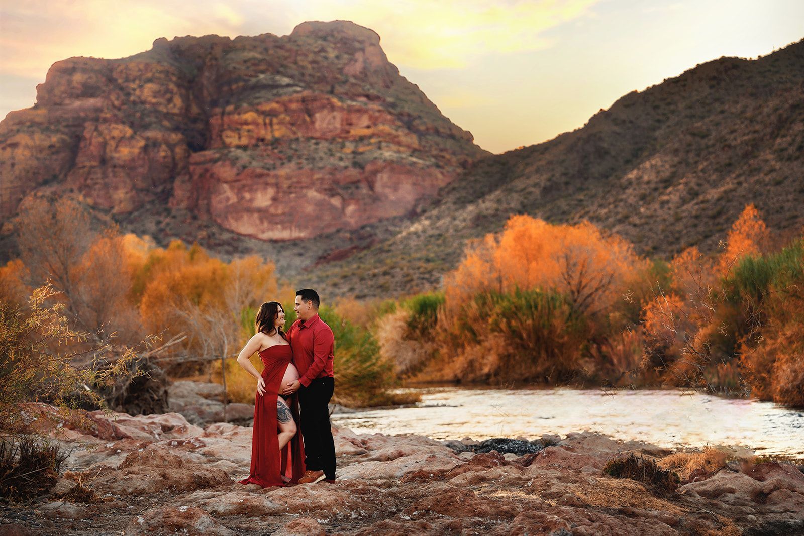 Couple in red attire standing by a river with desert cliffs and autumn trees at sunset.