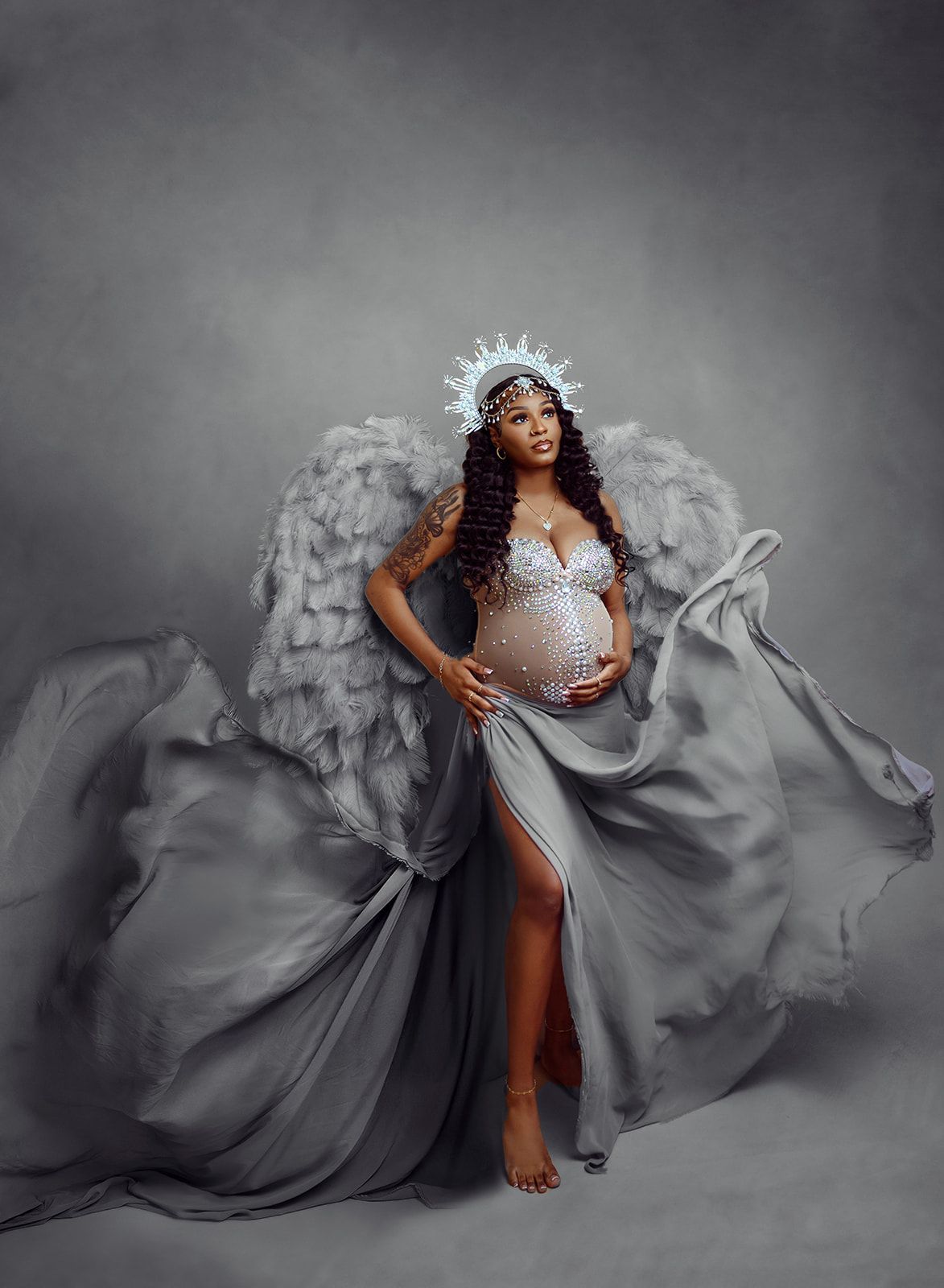 Pregnant model in a flowing gray gown and feathered cape, posing against a gray studio backdrop.