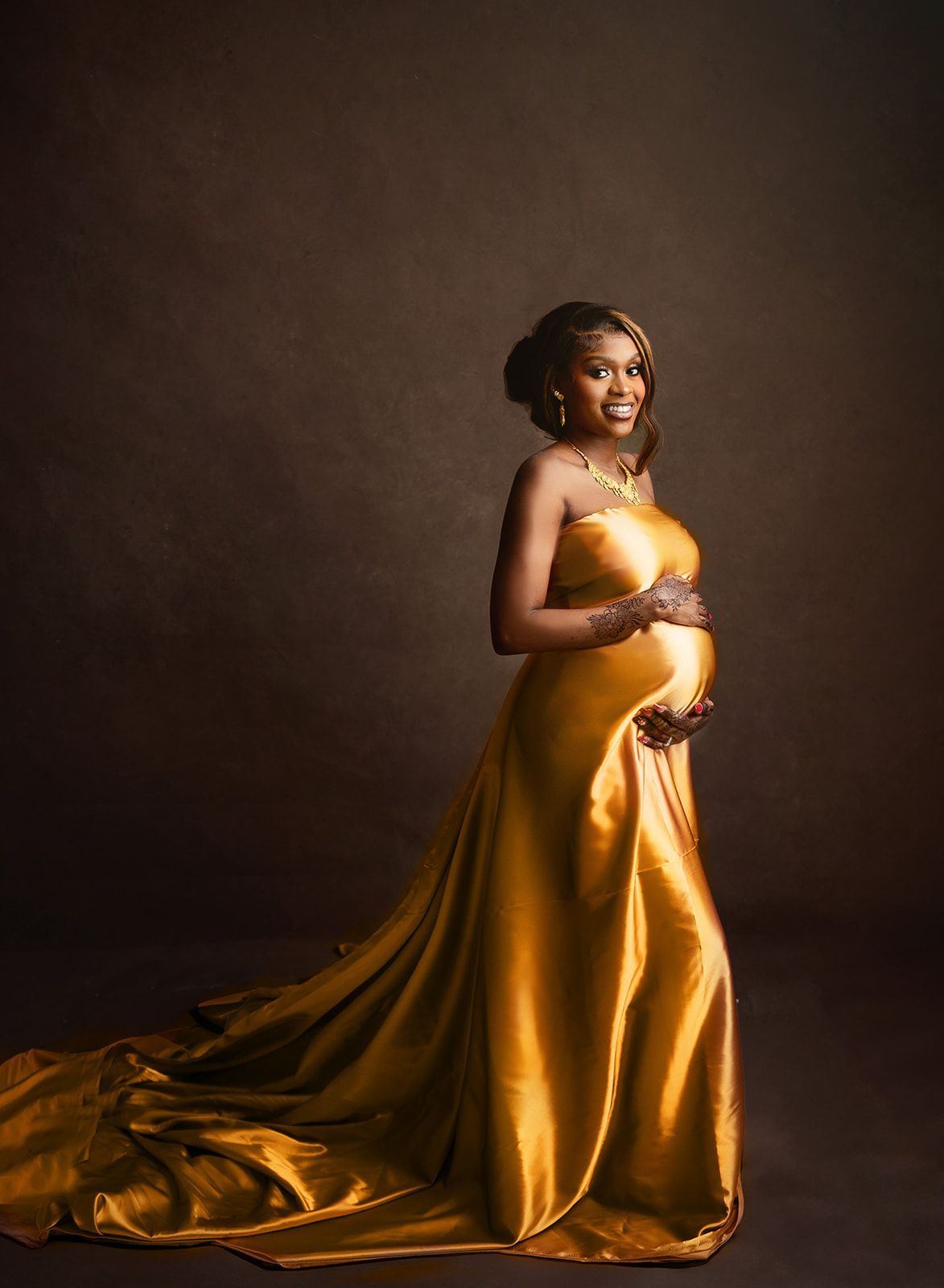 Pregnant woman in a gold satin gown posing against a dark studio backdrop.