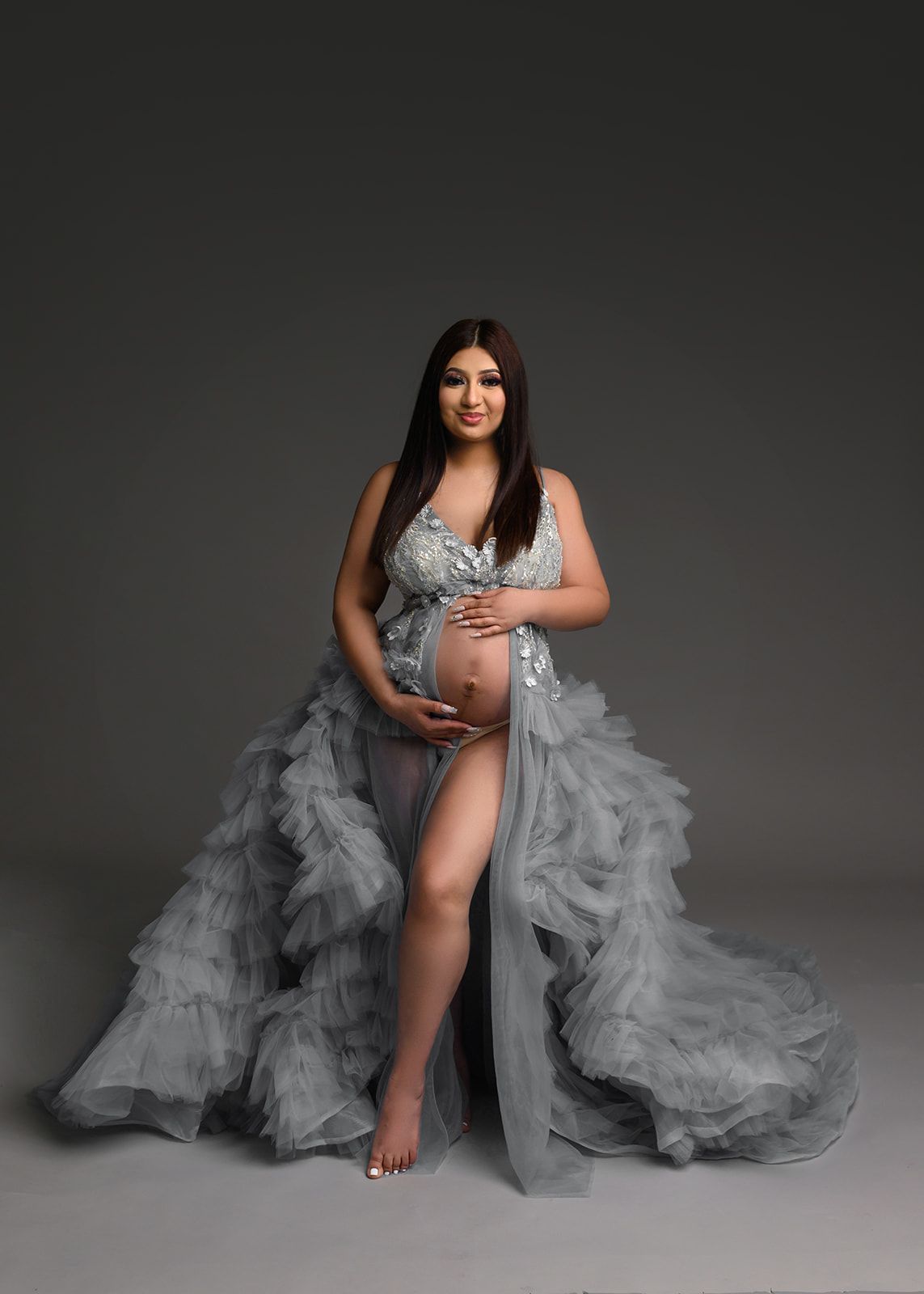 Pregnant woman in a silver gown with a flowing gray train, standing against a dark studio backdrop.