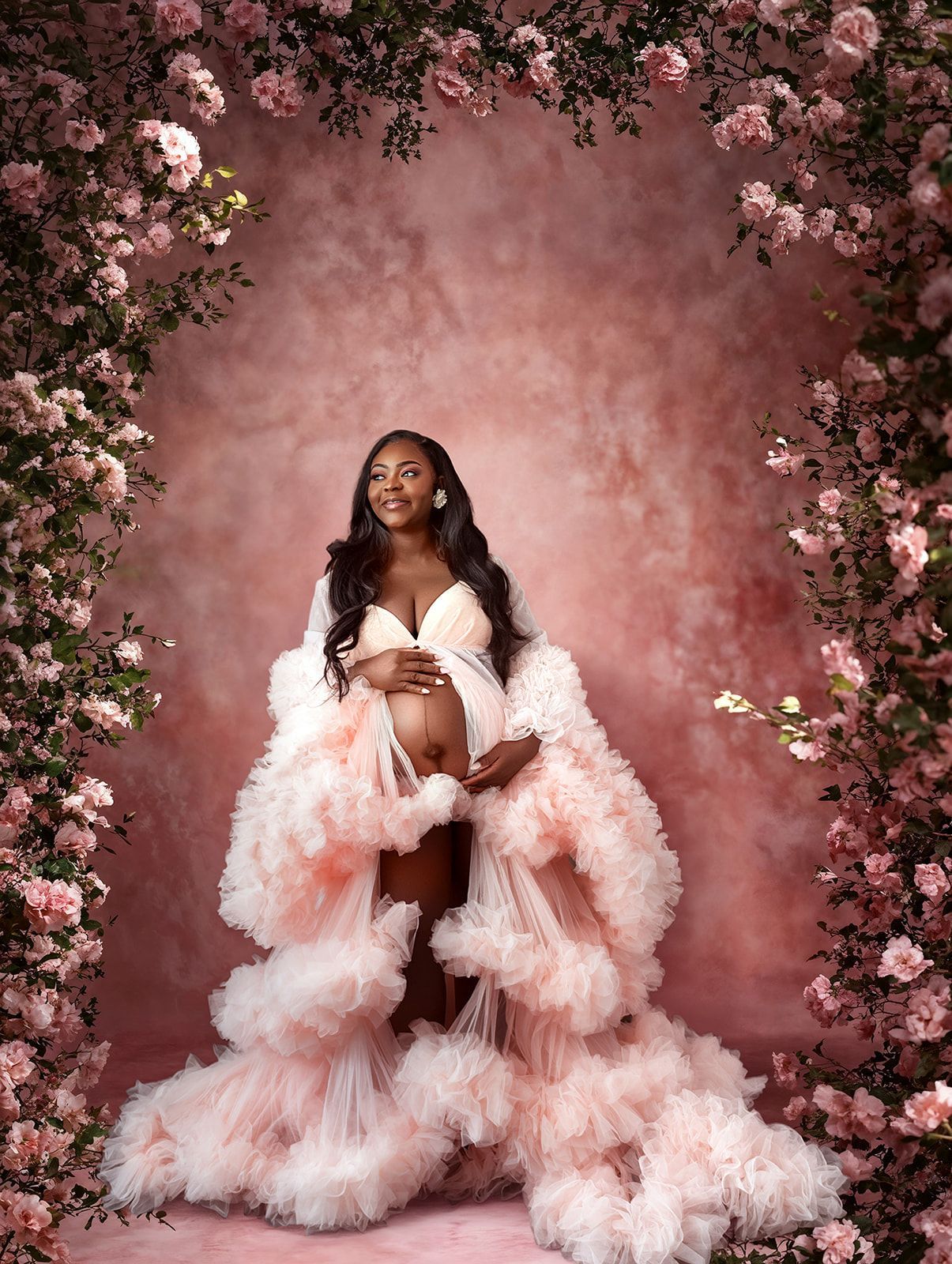 Pregnant person in a pink feathered gown against a floral pink backdrop.