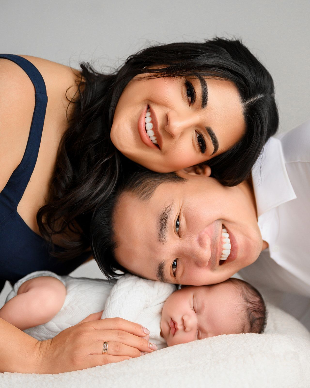 Smiling parents and sleeping newborn cuddled together on a bed.