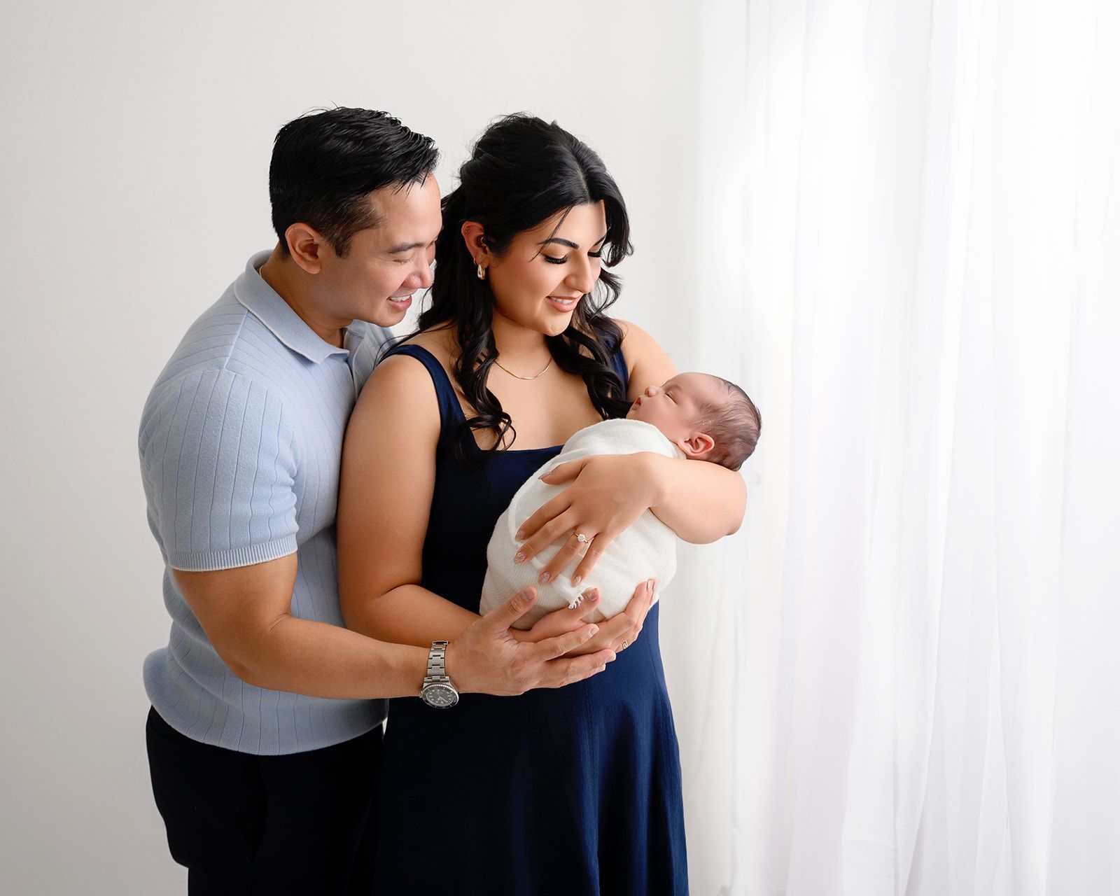 Parents gently holding a newborn by a white curtain, smiling softly in a bright studio.