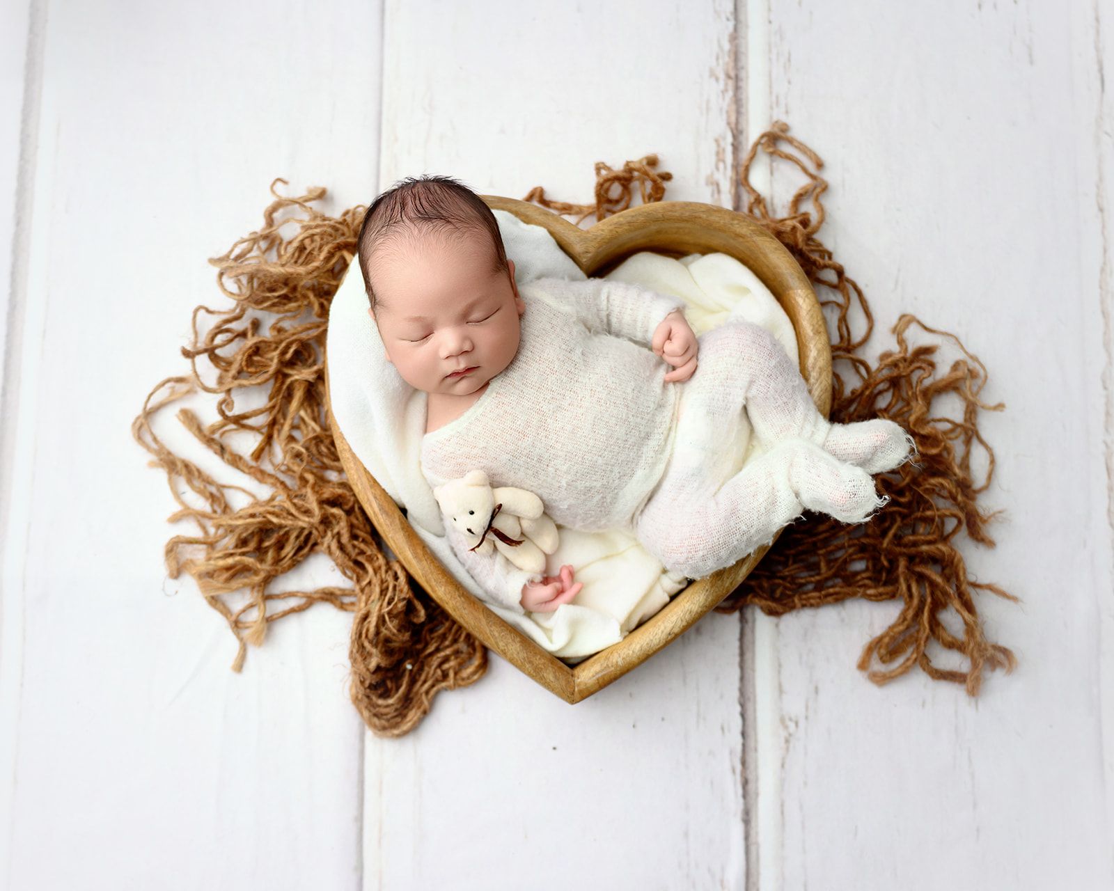 Sleeping baby in a white knit outfit on a heart-shaped cushion with a small teddy bear on a white wooden background.