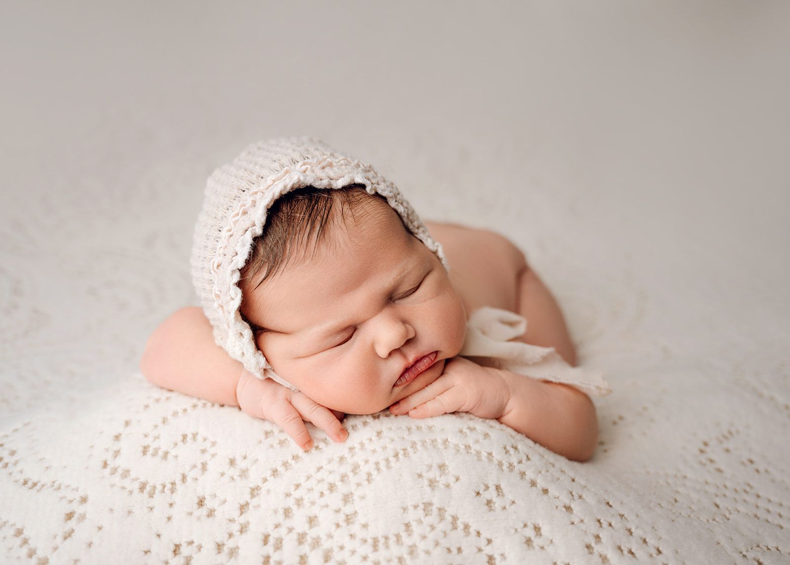 Sleeping newborn in a white knit bonnet, resting on soft white blankets with hands under chin.