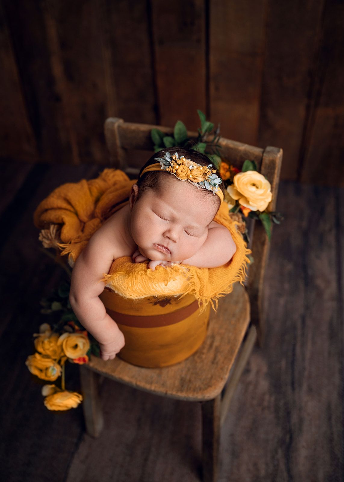 Sleeping baby in an orange wrap and floral headband, posed in a small chair with a rustic wooden backdrop.