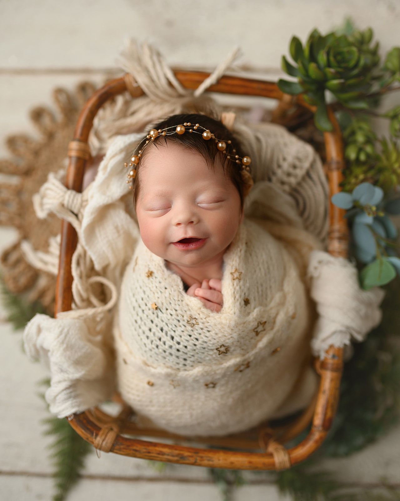 Sleeping newborn wrapped in cream knit, lying in a wicker basket with greenery and white flowers.