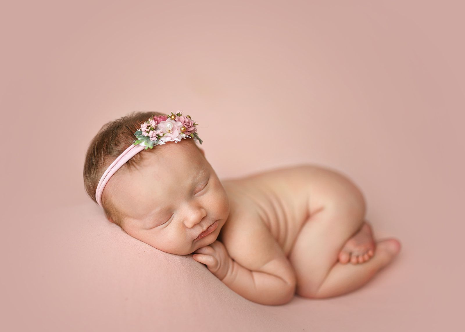 Sleeping baby curled on a pink blanket, wearing a floral headband.