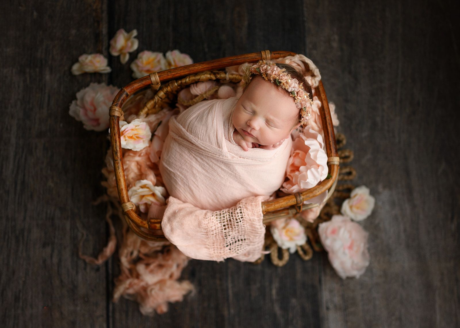 Sleeping baby in a pink blanket nestled in a small basket with flowers on a dark wooden background.