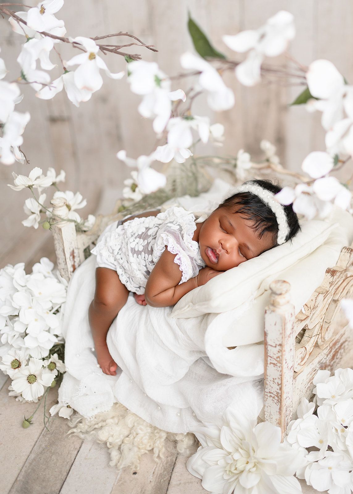 Sleeping newborn on a white cushioned bed surrounded by soft white flowers and lace decorations.