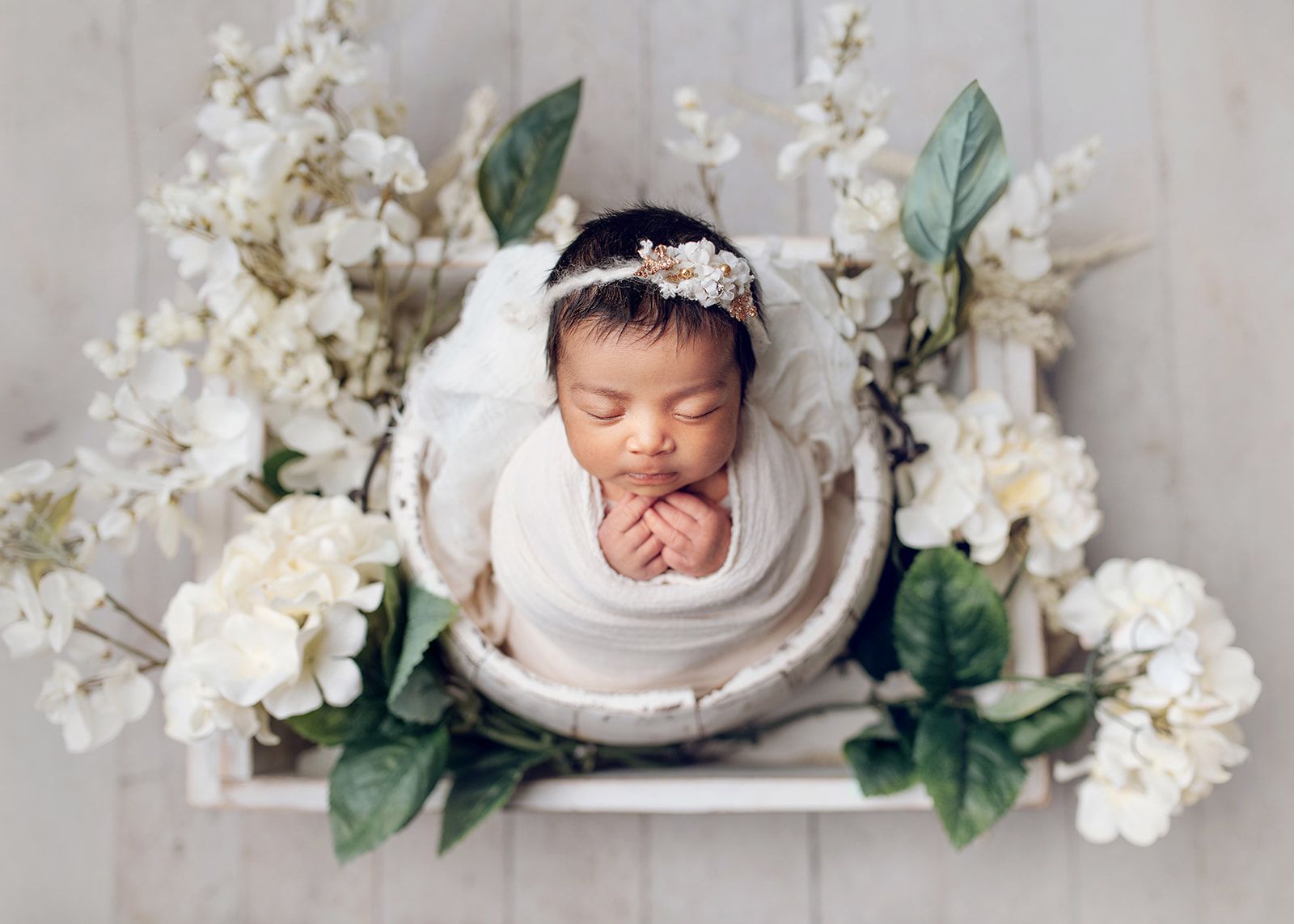 Sleeping newborn wrapped in white, surrounded by white flowers and green leaves.