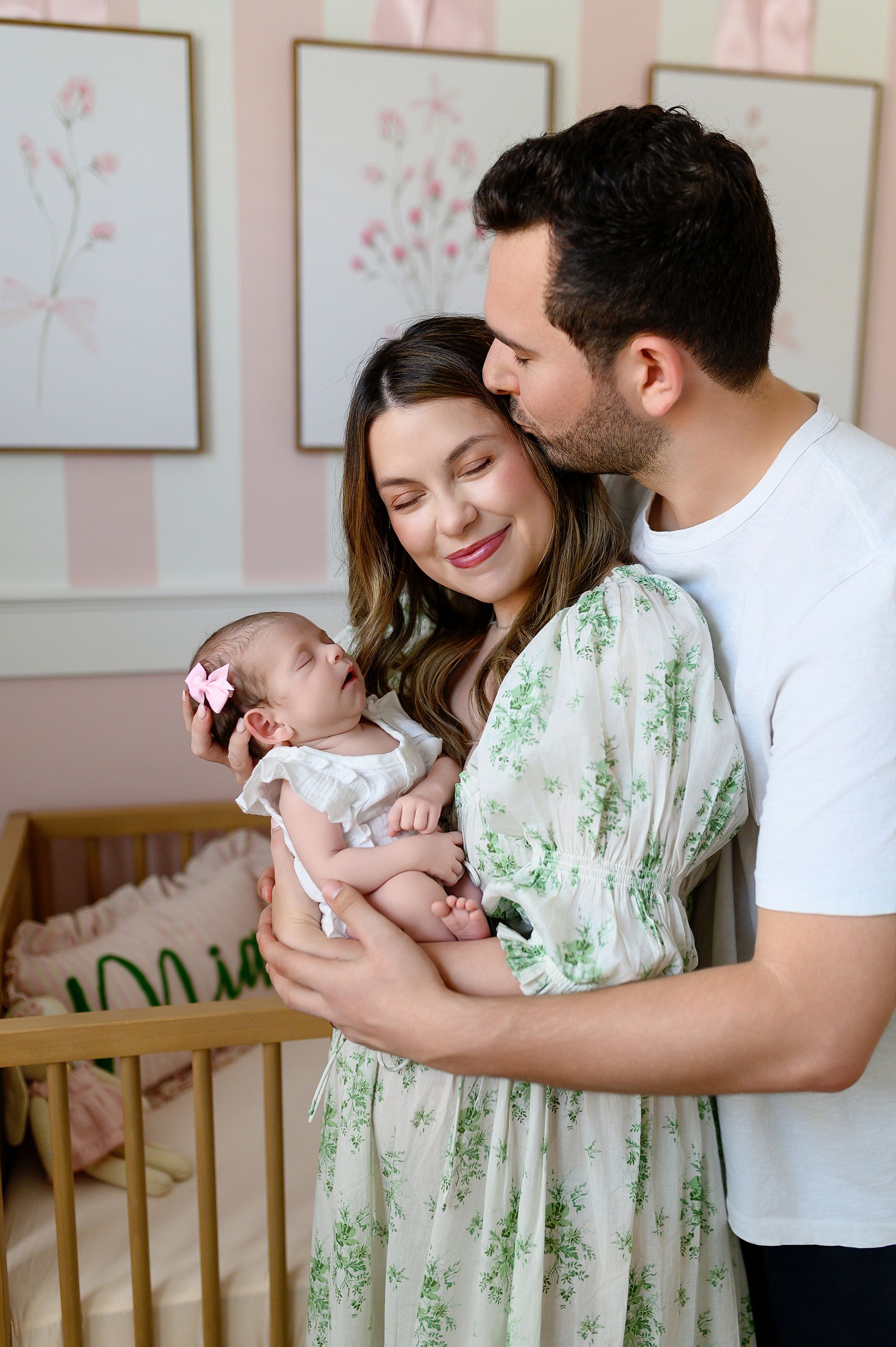 Couple gently holding a baby in a pastel nursery, with framed floral art on the wall.