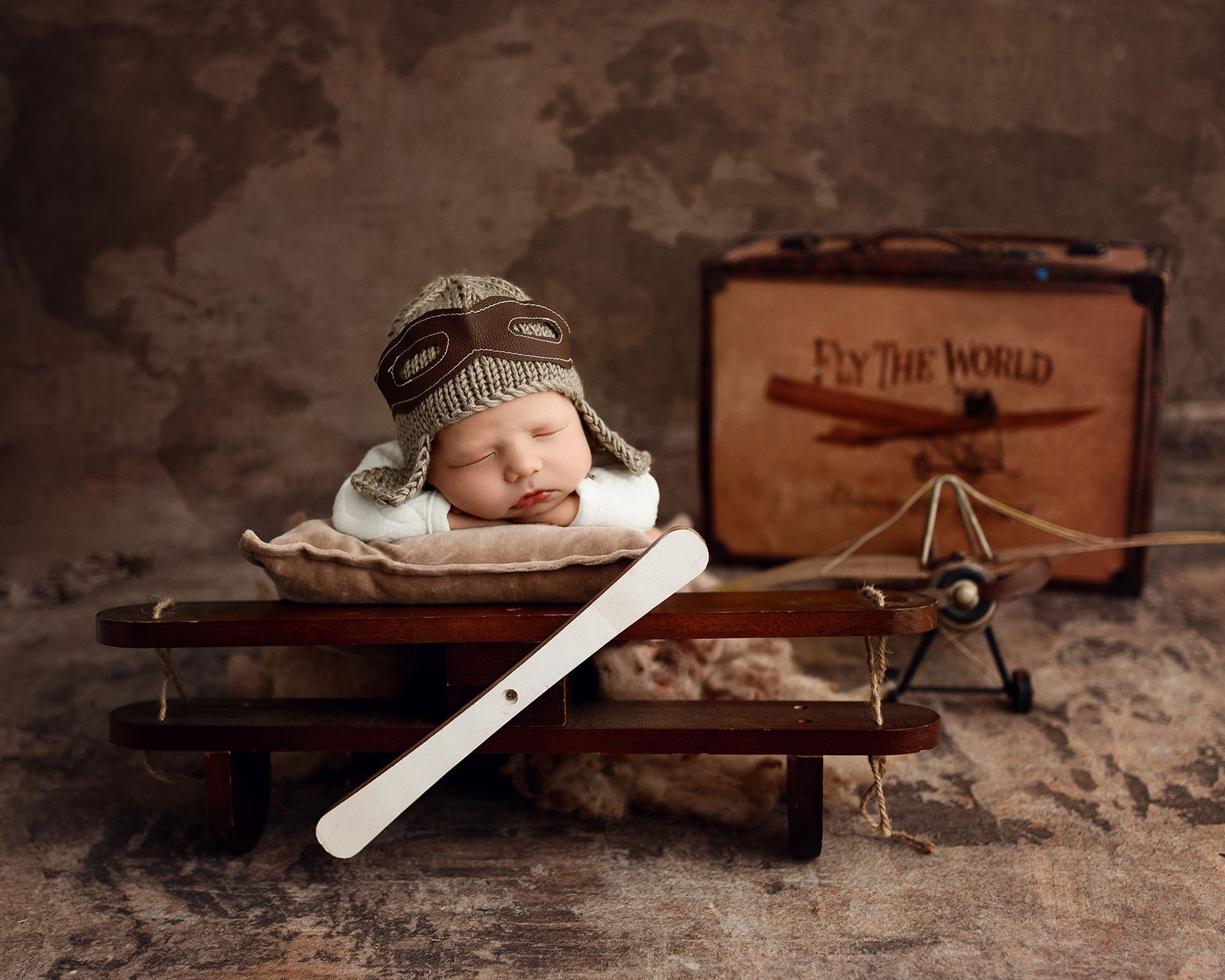 Sleeping baby in a knit hat on a wooden sled beside a suitcase labeled “Fly the World.”