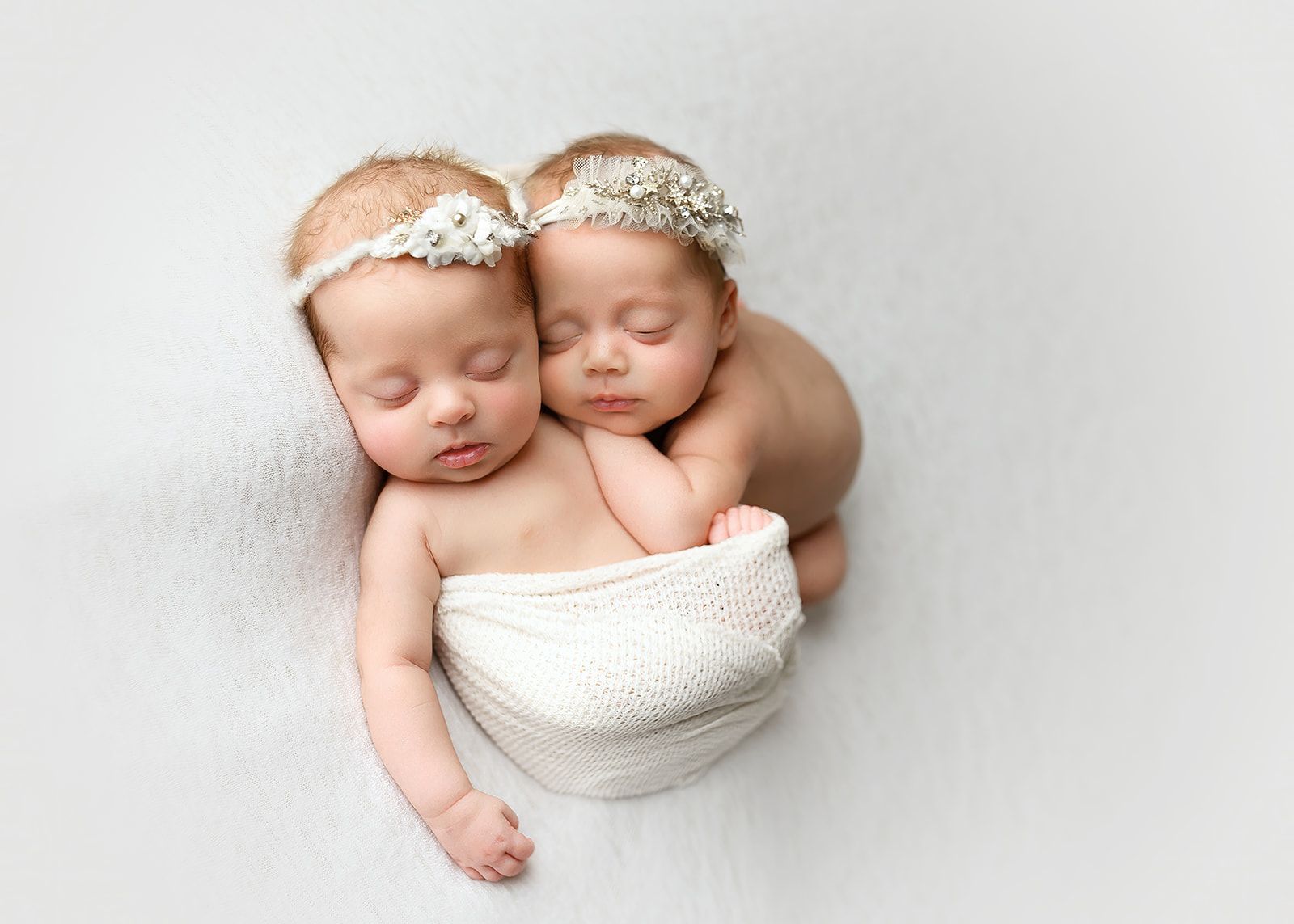 Two sleeping babies cuddled together in a white blanket with floral headbands on a light background.