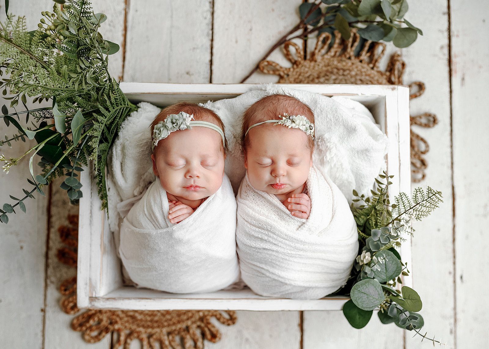 Two sleeping newborns swaddled in white blankets on a rustic wooden tray with greenery.
