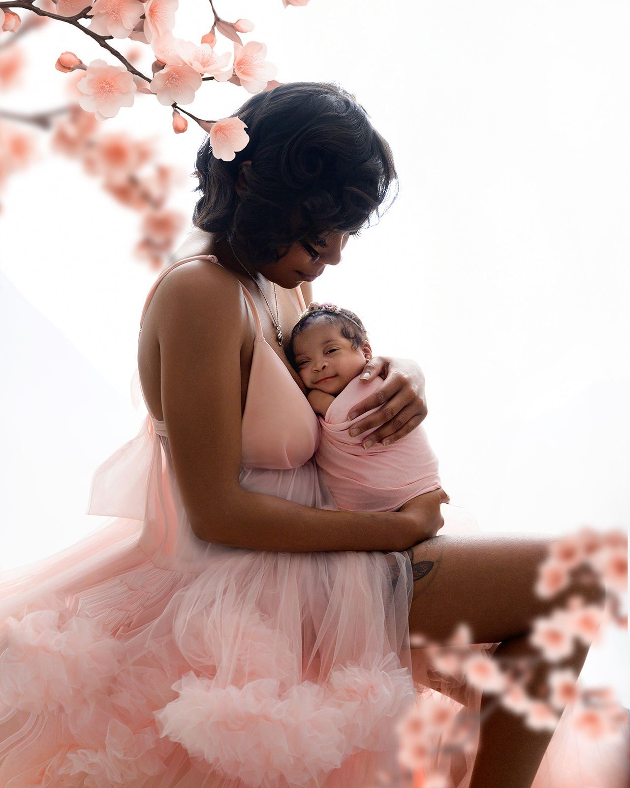 Woman in a pink tulle dress gently holding a baby, with soft floral decor in the background.