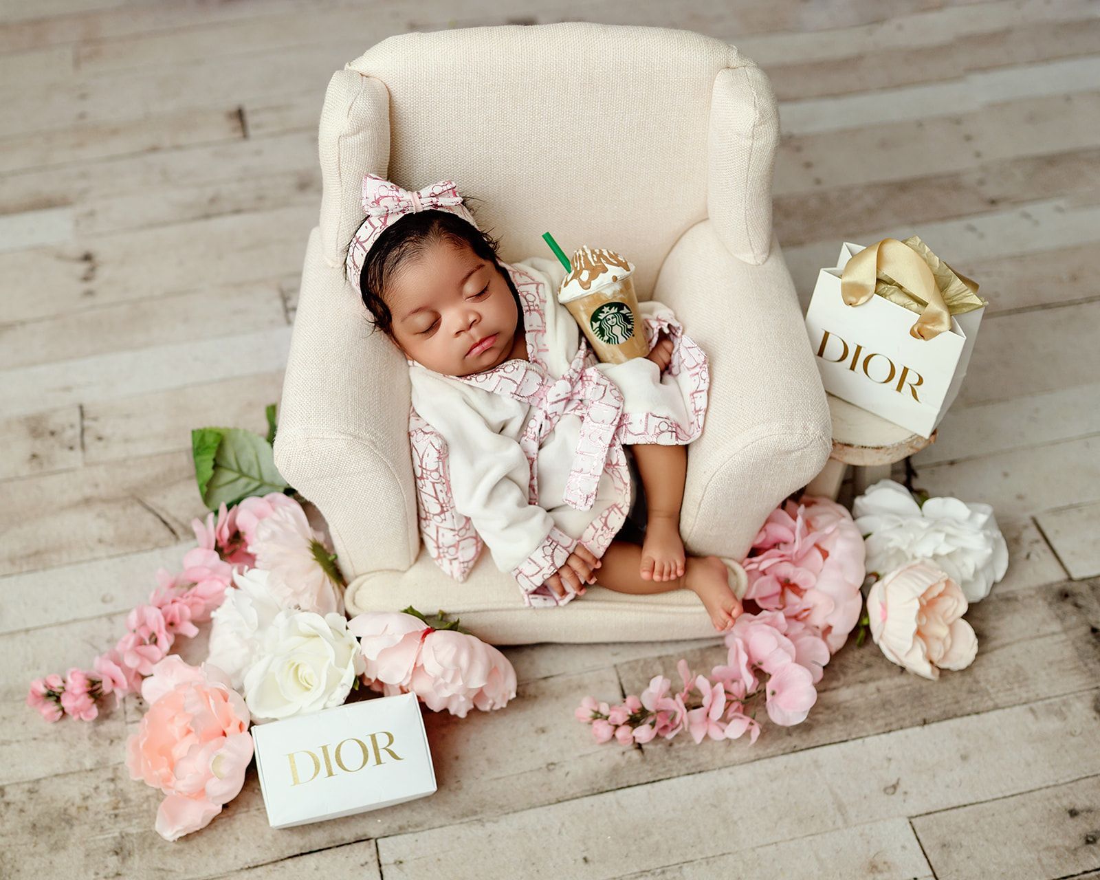 Sleeping newborn in a white chair, surrounded by pink flowers and Dior gift boxes on a light wood floor.