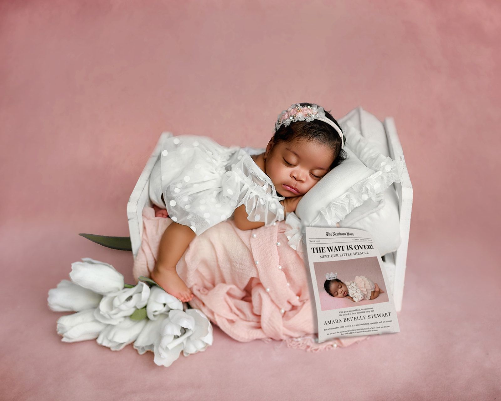 Sleeping baby in a white outfit on pink fabric, beside a soft blanket and a small card.