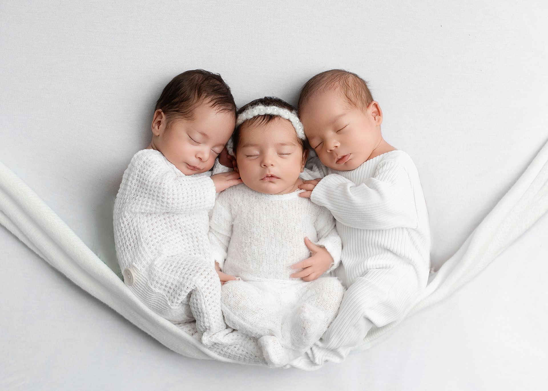 Three sleeping newborns in white blankets nestled together in a soft white hammock.