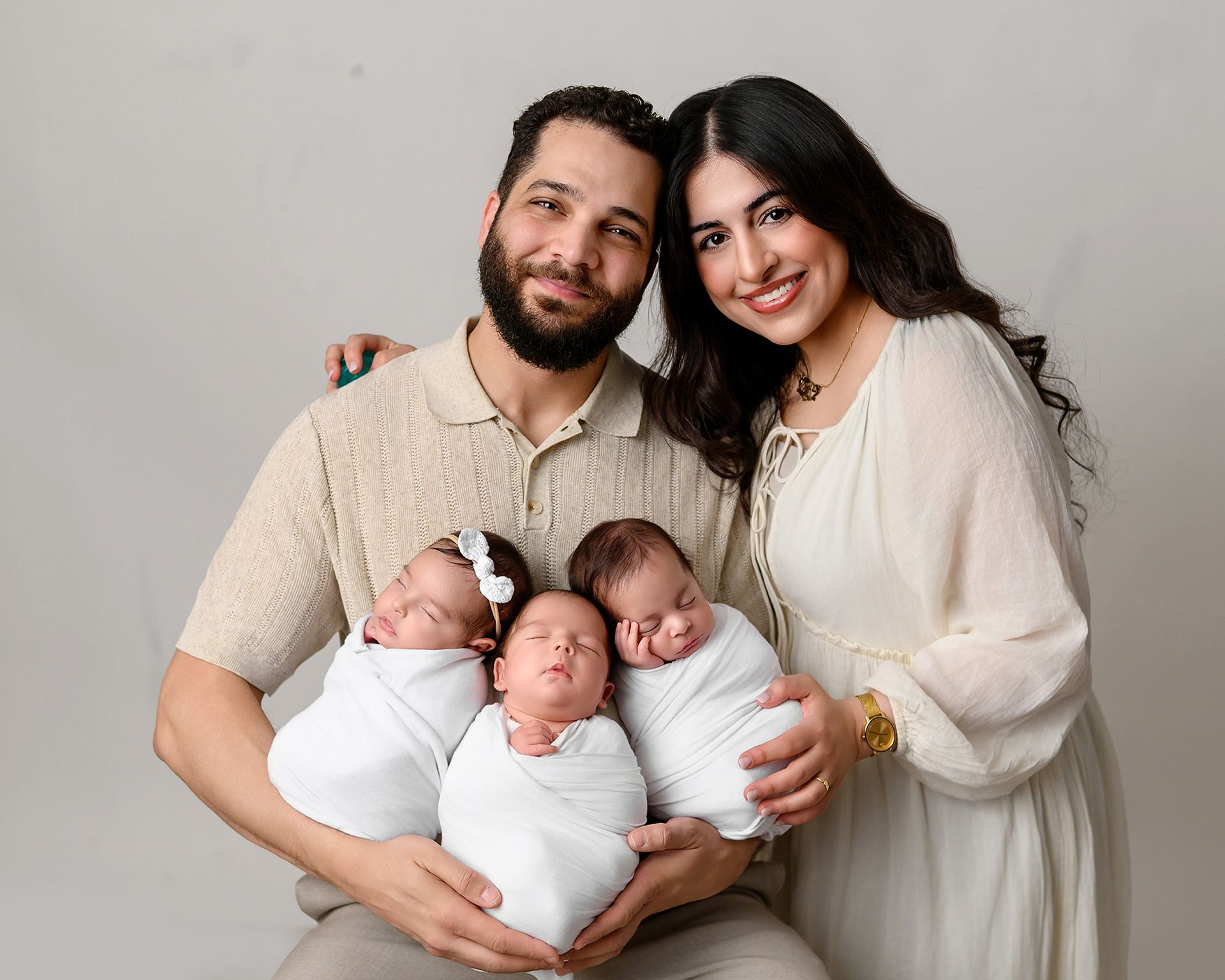 A smiling family portrait with two adults holding two swaddled newborns against a plain beige background.