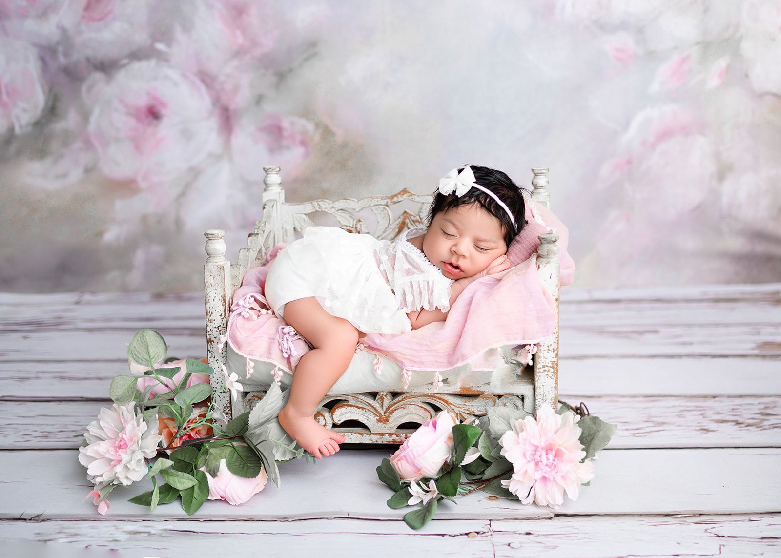 Sleeping baby on a pink cushioned bed surrounded by flowers against a soft pastel backdrop.