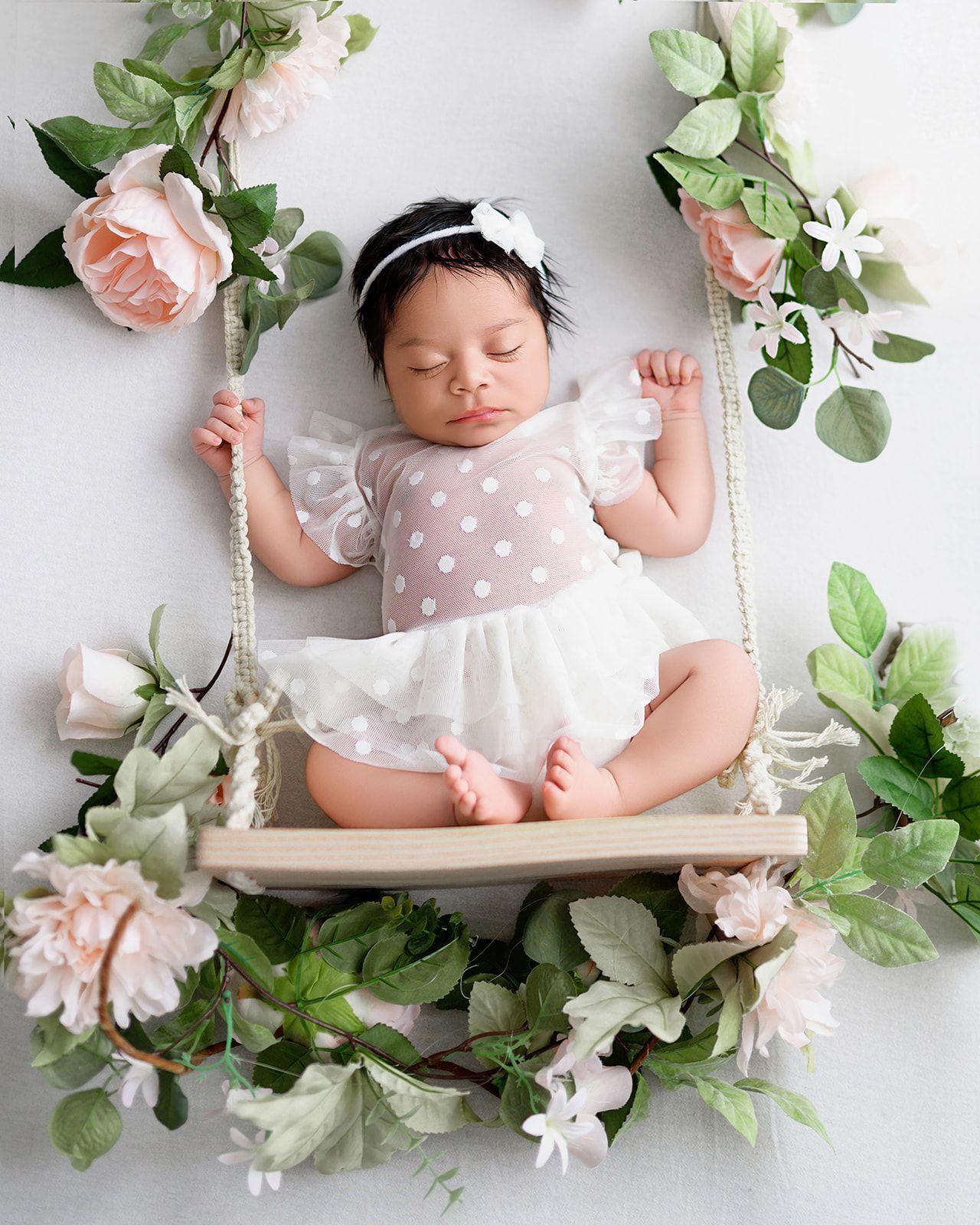 Sleeping baby in a pink outfit on a floral swing with greenery and white flowers.