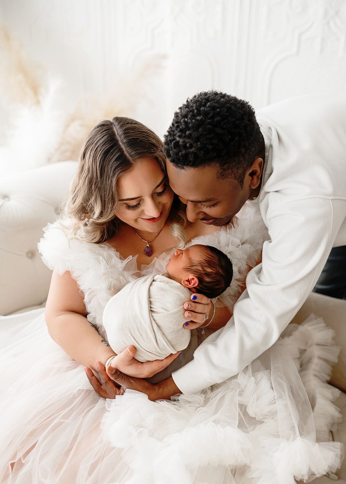 New parents in white clothing gently cradling a swaddled newborn on a bed.