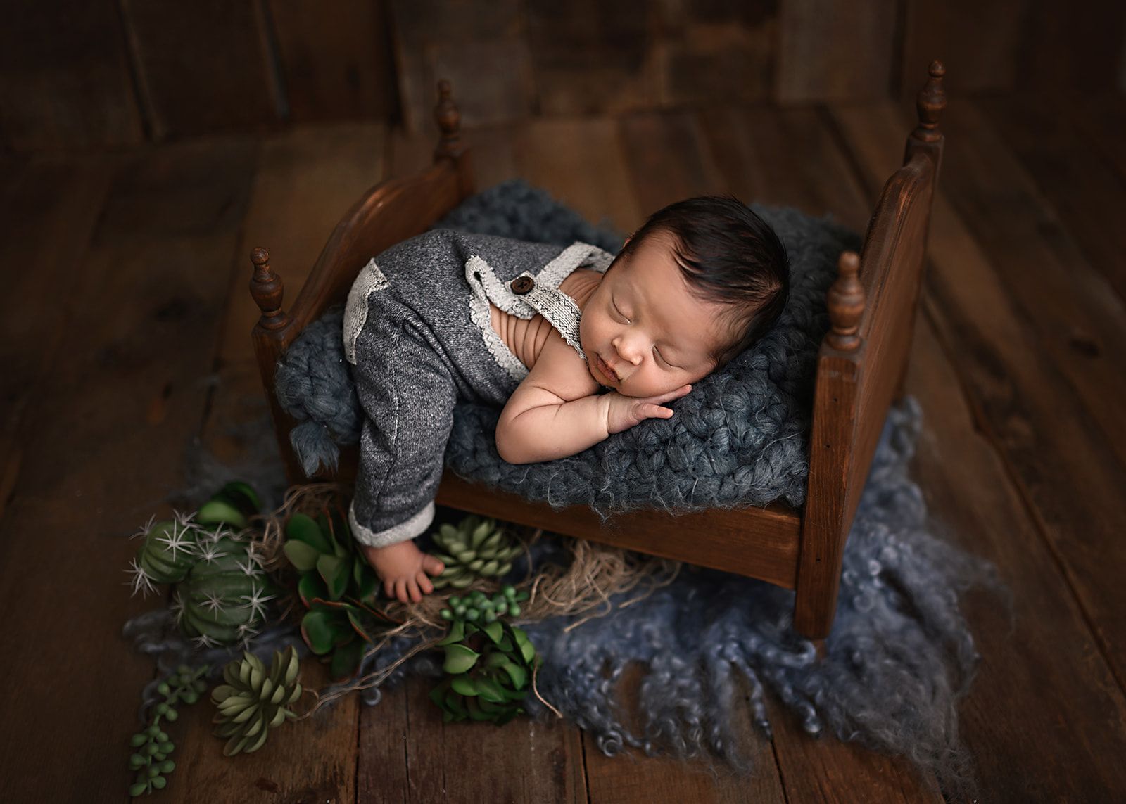 Sleeping baby on a wooden chair with a gray blanket, surrounded by greenery on a rustic floor.