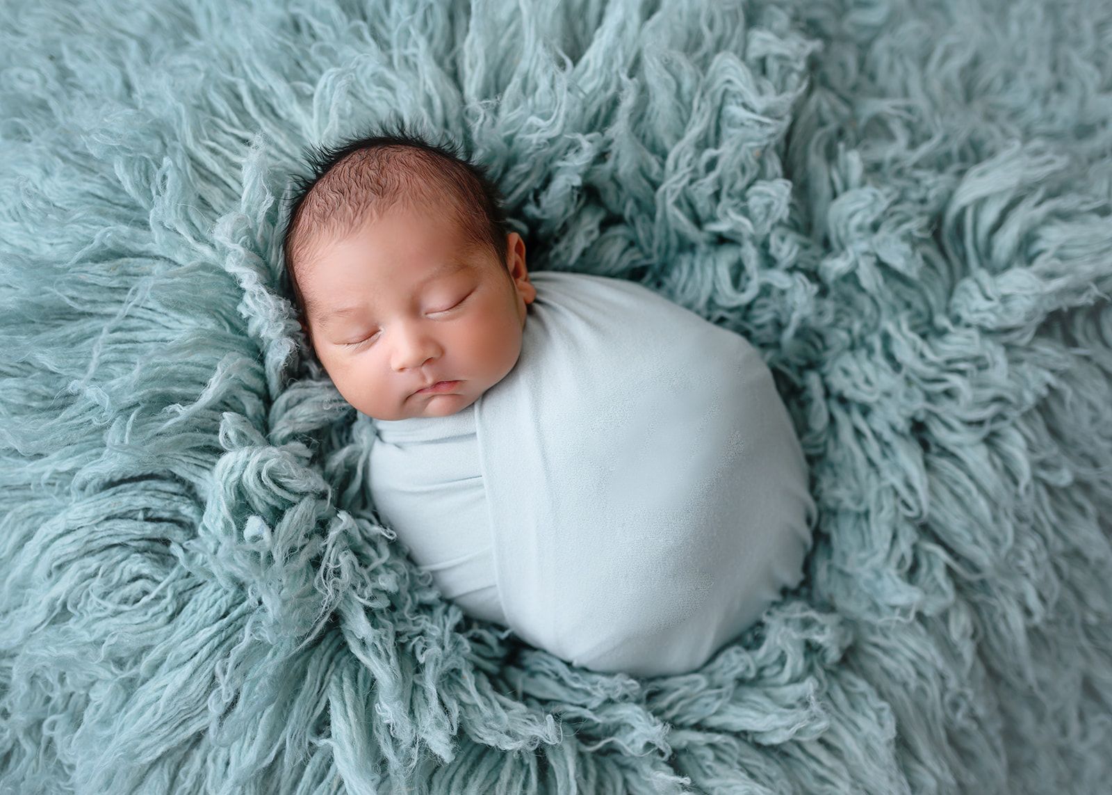 Sleeping newborn swaddled in pale blue on a fluffy gray blanket.