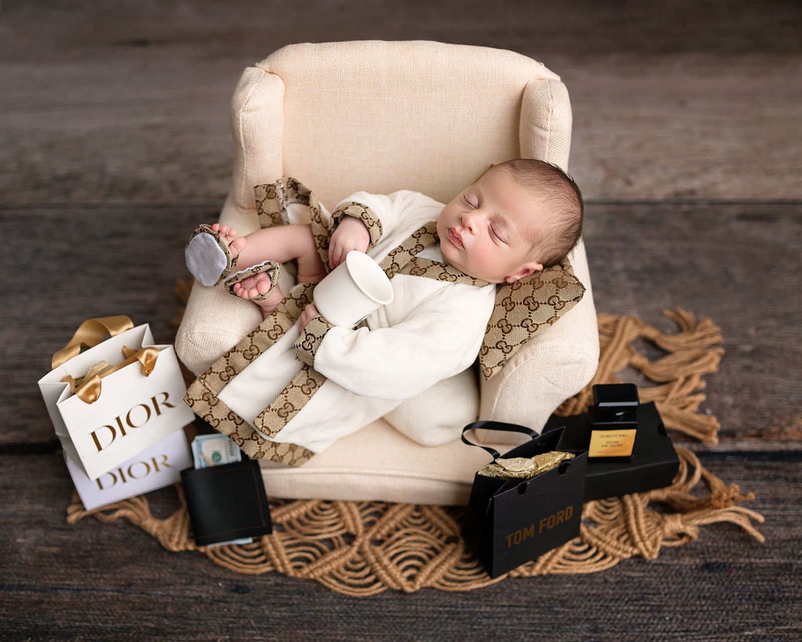 Sleeping baby in a cream chair with DIOR gift bags, wrapped in white on a woven mat against a wooden backdrop.