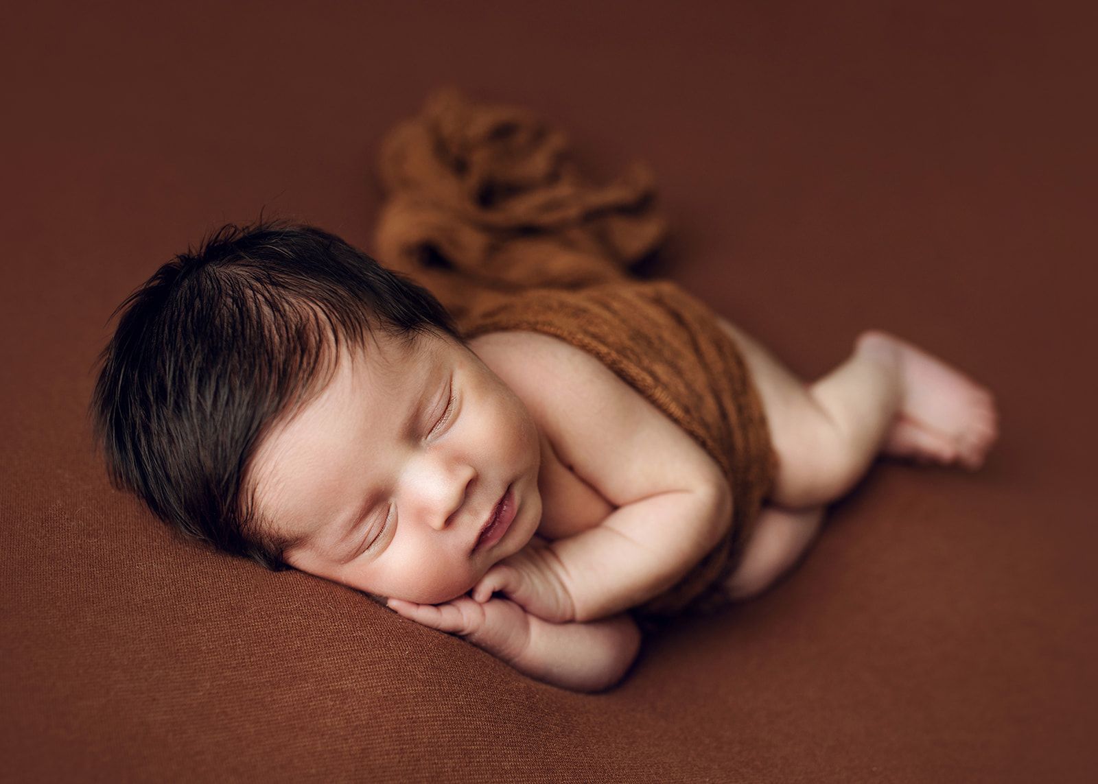 Sleeping newborn wrapped in brown fabric on a soft brown background.