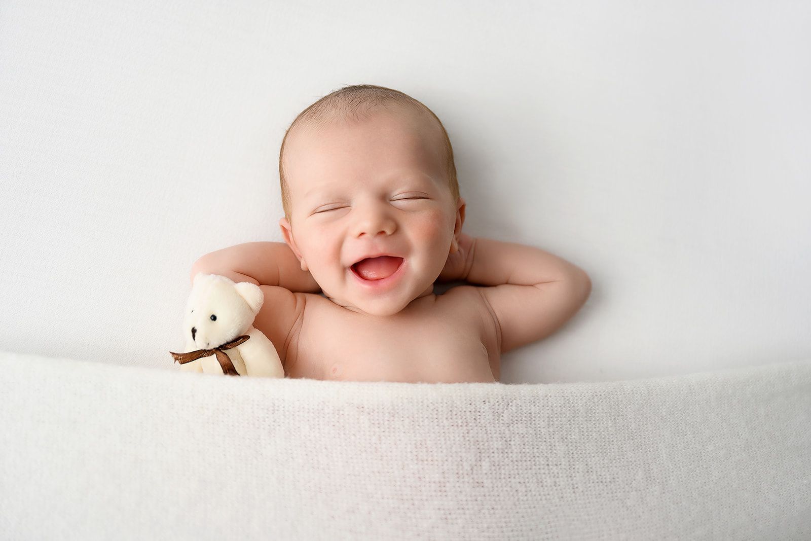 Sleeping baby in a white tub, smiling with arms raised, holding a small teddy bear.