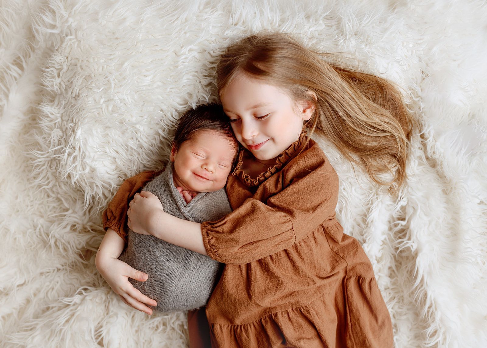 Sleeping baby and smiling child lying together on a fluffy cream blanket, wrapped in soft neutral clothing.