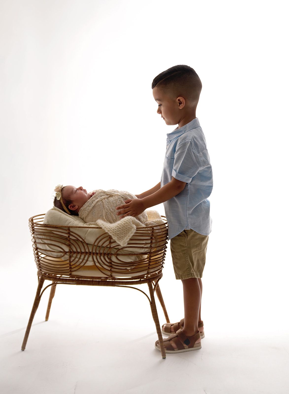 Young child standing beside a wicker bassinet, gently touching a sleeping baby inside.