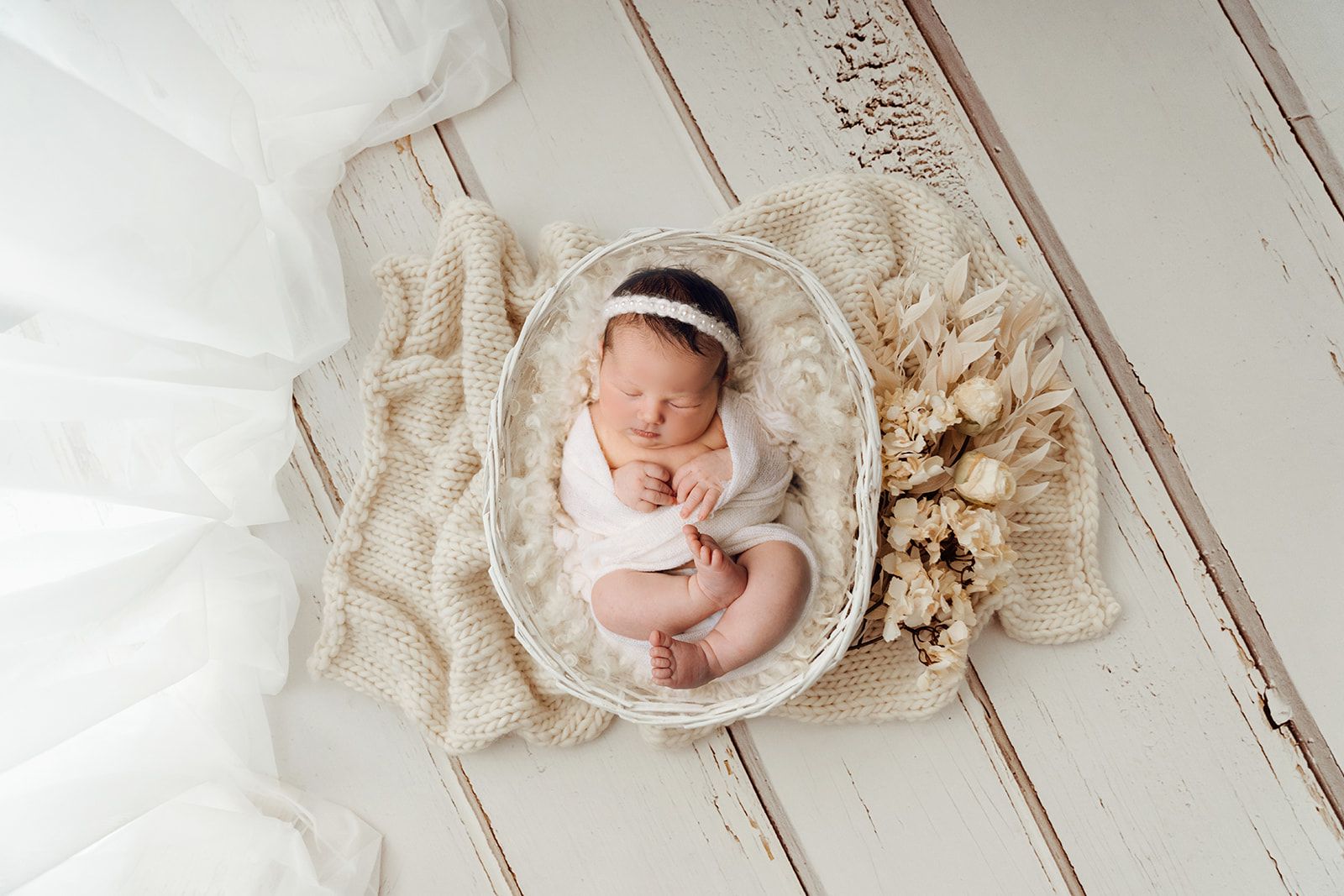 Sleeping newborn in a cream knit basket nest on a soft white blanket, surrounded by dried florals.