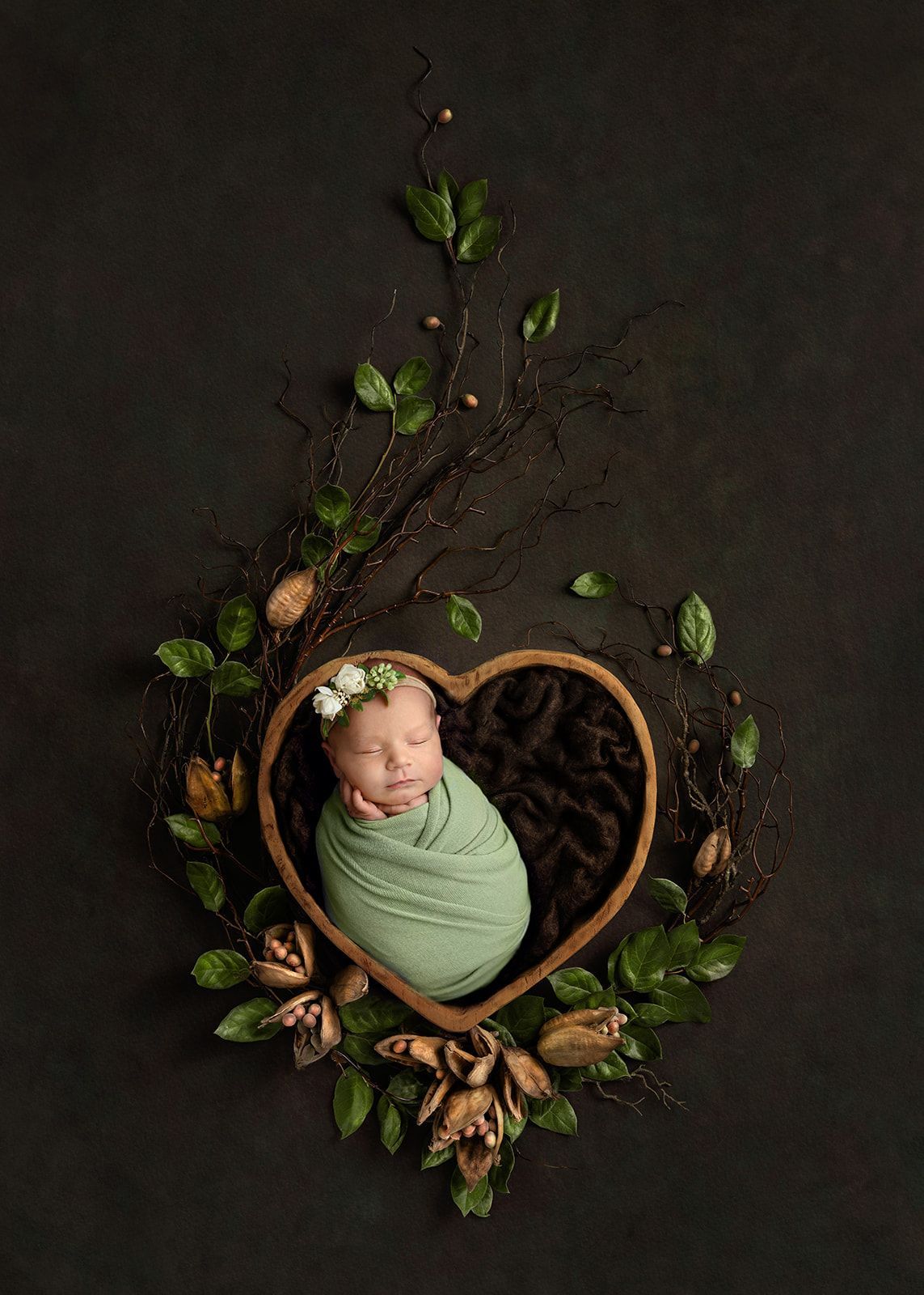 Sleeping baby wrapped in green swaddle inside a heart-shaped wicker nest with leaves on a dark background.