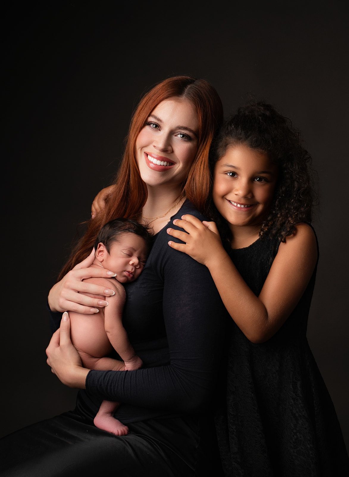 Smiling woman holding a sleeping baby, with a young girl hugging her in a dark studio portrait.