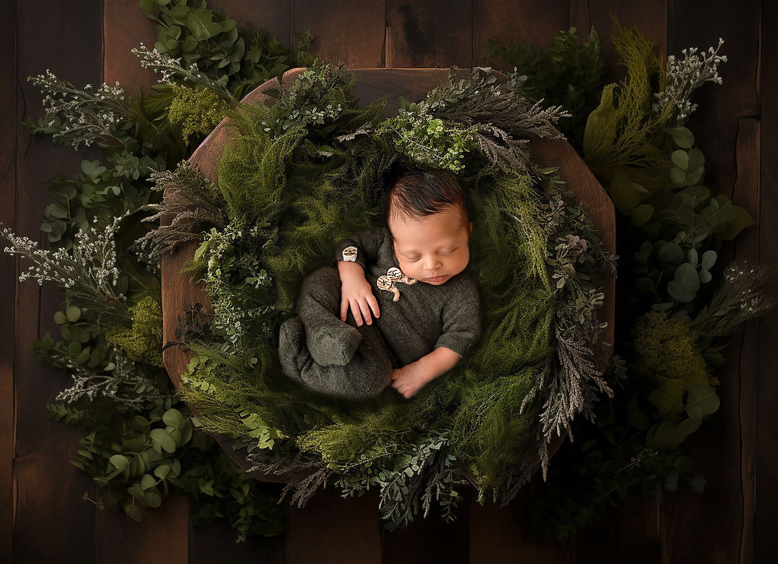 Sleeping baby nestled in a wreath of greenery on a dark wooden background.