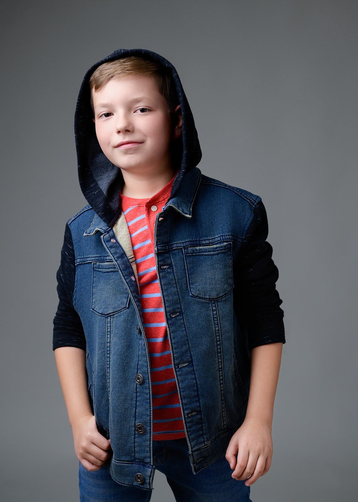 Person in a dark denim vest with hood over a red striped shirt, standing against a gray background.