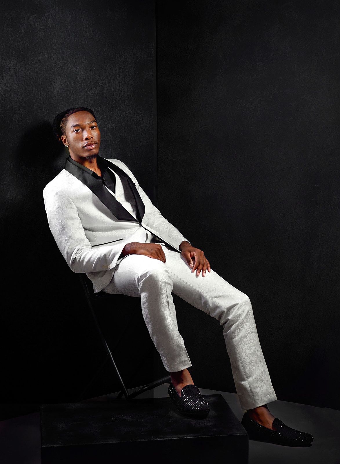 Man in a white suit seated on a chair against a dark background.