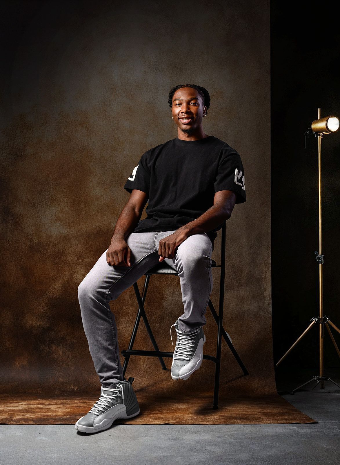 Man seated on a folding stool in a studio, wearing a black T-shirt, gray pants, and white sneakers, with a light stand nearby.